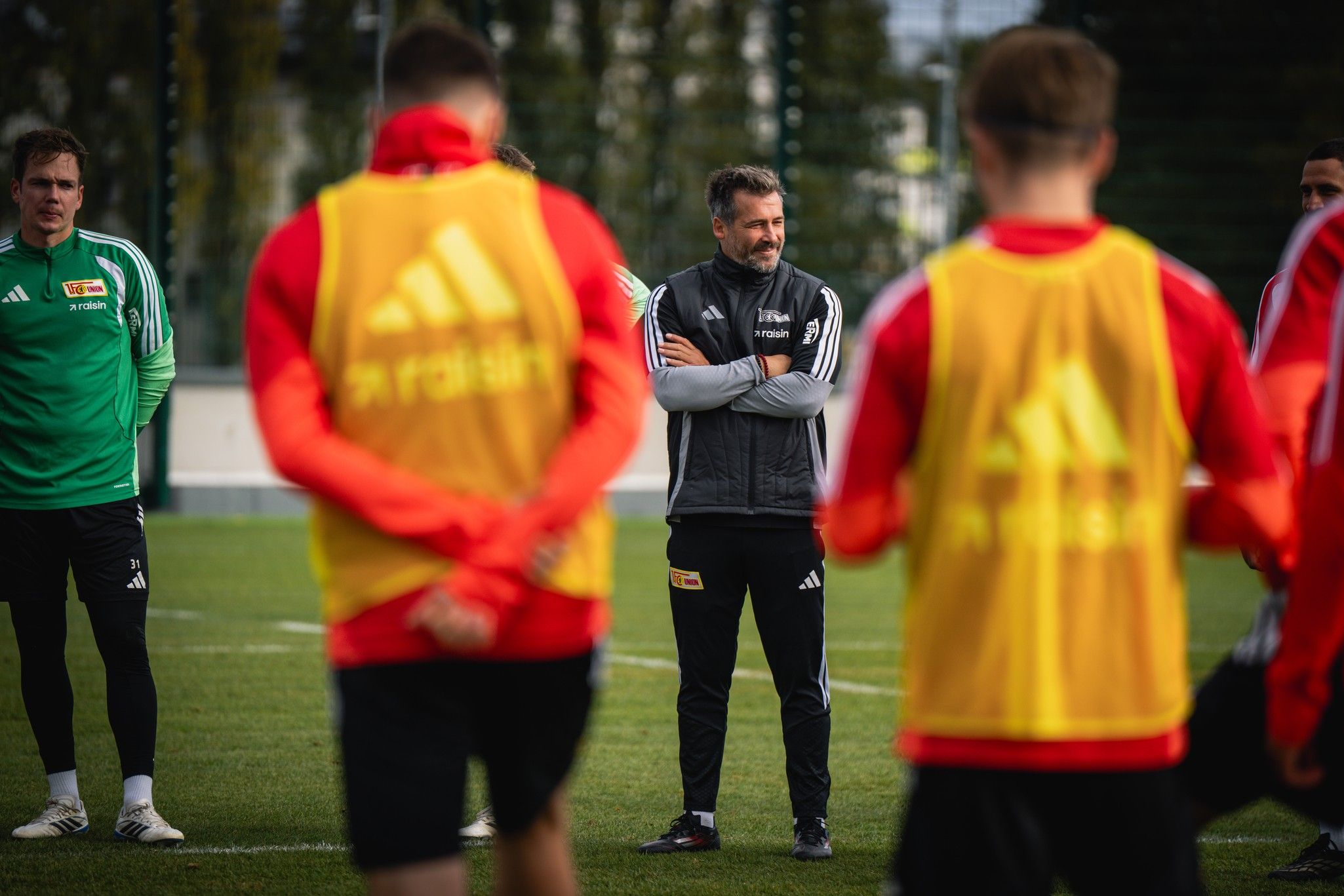 Ein Fußballtrainer steht mit verschränkten Armen im Vordergrund, während Spieler in Trainingswesten ihm zuhören.