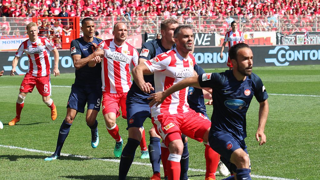 Fußballspieler stehen in einer spannenden Spielsituation auf dem Platz, umgeben von jubelnden Fans im Hintergrund.