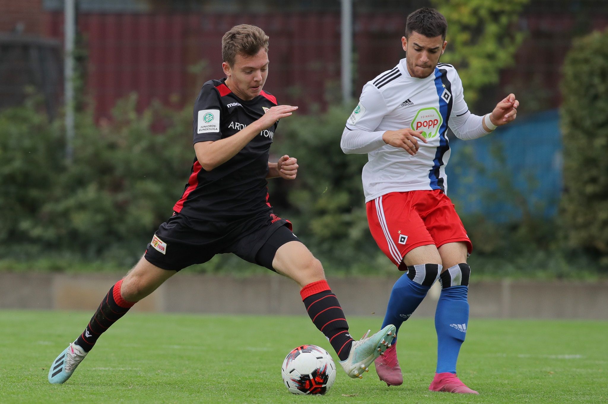 Zwei Fußballspieler kämpfen um den Ball auf einem grünen Rasenfeld. Der Spieler in Schwarz hat einen angreifenden Blick.