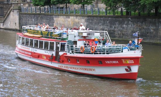 Ein rotes Passagierschiff mit dem Namen "Viktoria" fährt auf einem ruhigen Gewässer, umgeben von Bäumen und Uferpromenade.