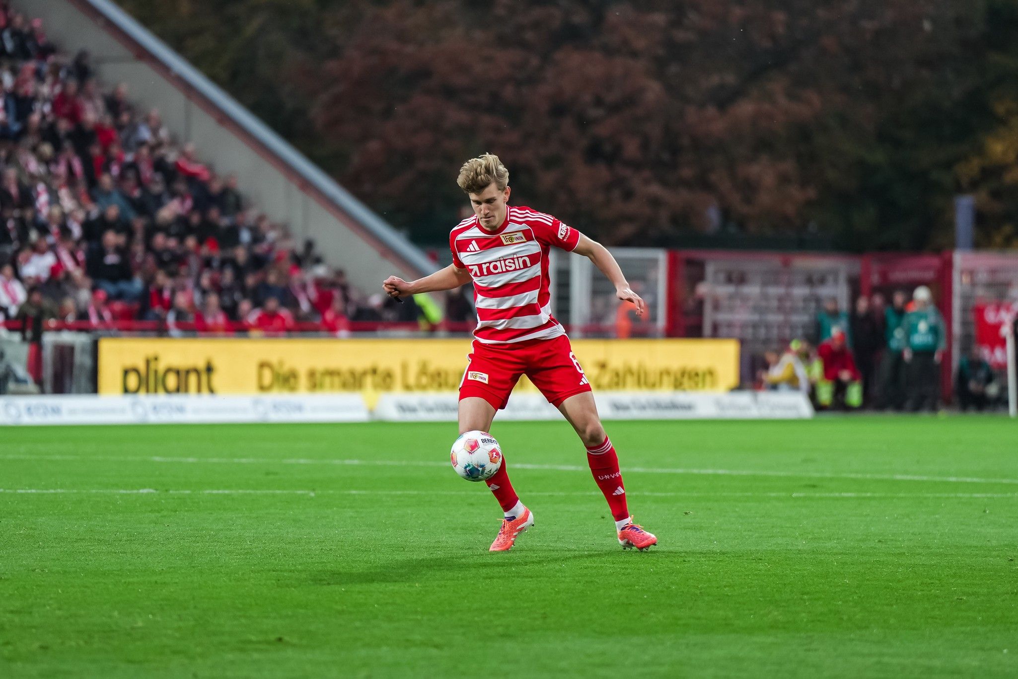 Fußballspieler in rot-weißer Uniform kontrolliert den Ball auf einem Stadionrasen. Fans im Hintergrund.