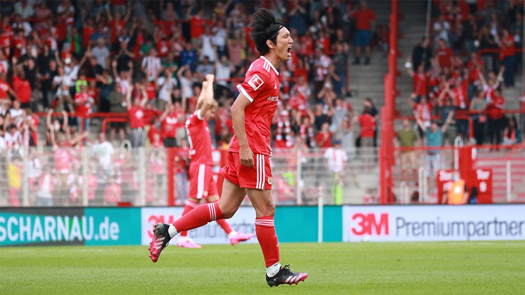 A player in a red jersey celebrates after a goal, surrounded by cheering fans in the stadium.