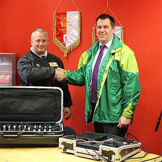 Zwei Männer schütteln sich die Hände vor rotem Hintergrund, während Ausrüstungen und ein Koffer auf dem Tisch stehen.