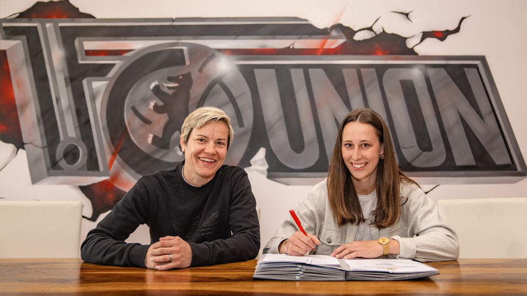 Two people are sitting at a table, one of them is signing a contract. A graphic logo is visible in the background.