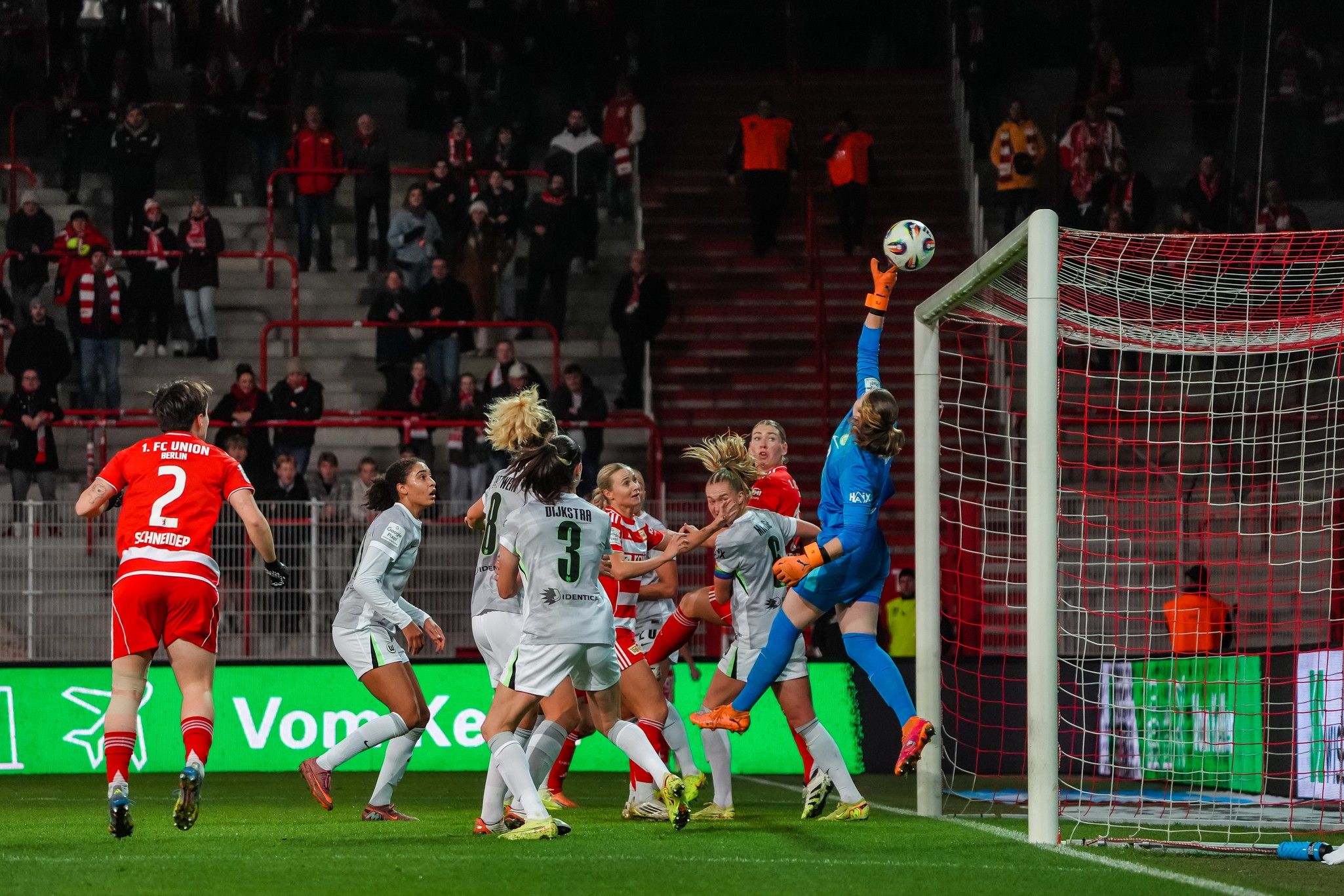 Eine Torhüterin springt, um einen Ball zu fangen, während Spielerinnen auf dem Feld um den Ball kämpfen. Stadionatmosphäre im Hintergrund.