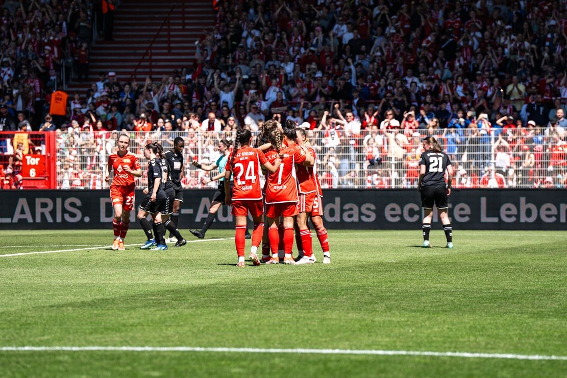 Spieler in roten Trikots feiern ein Tor, umgeben von jubelnden Fans im Stadion.