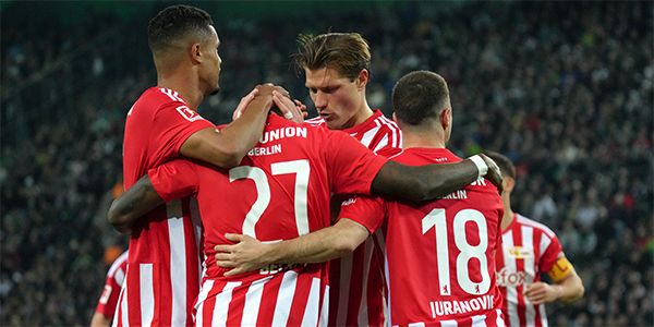 Players of the team celebrate in red-and-white jerseys, hugging after a goal in a stadium.