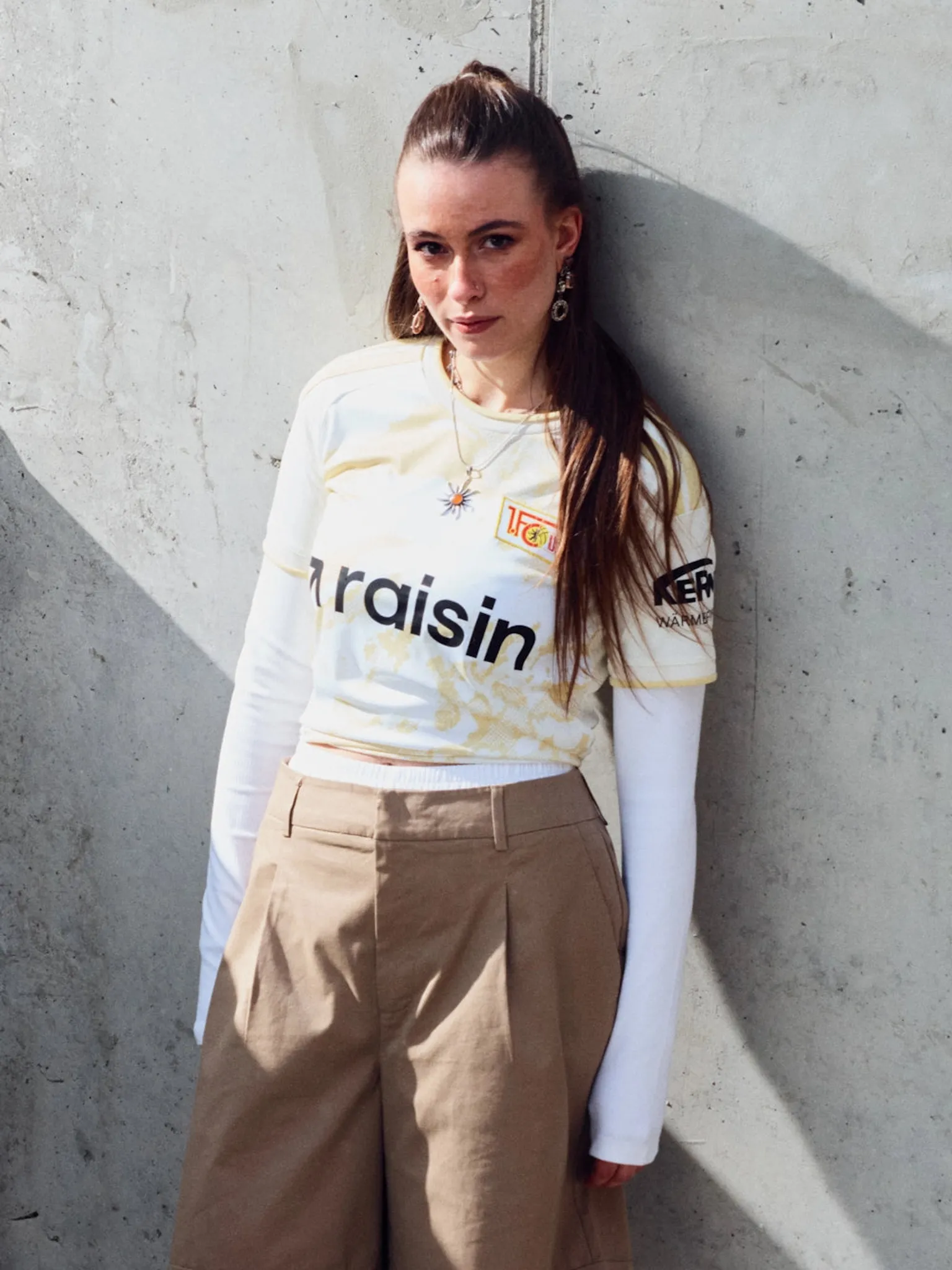 Eine Person in einem Fußballtrikot mit langen Ärmeln und weiten Hosen steht vor einer grauen Wand.