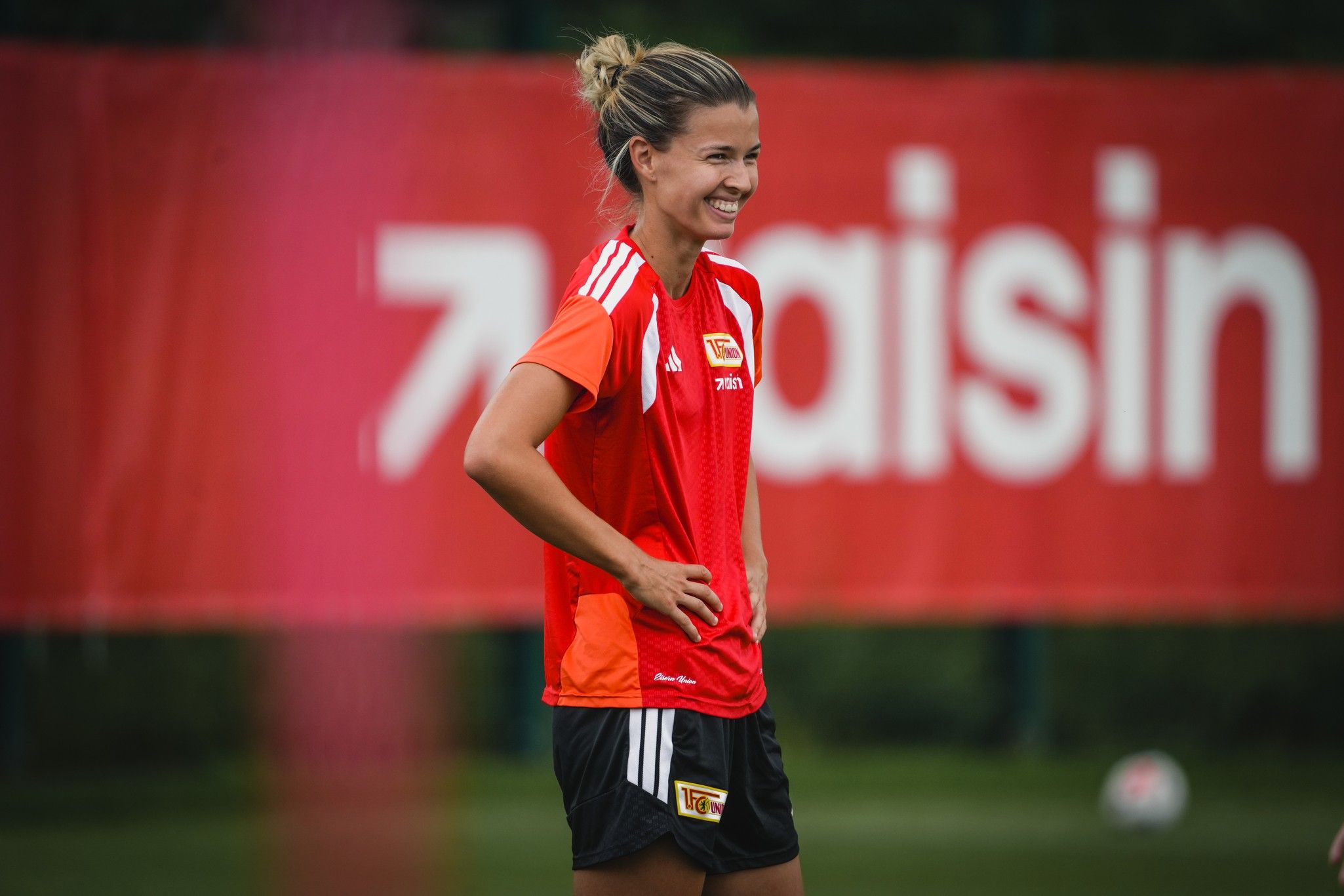 Eine lächelnde Fußballspielerin in rotem Trikot steht auf dem Trainingsplatz vor einem Banner mit der Aufschrift "aisin".