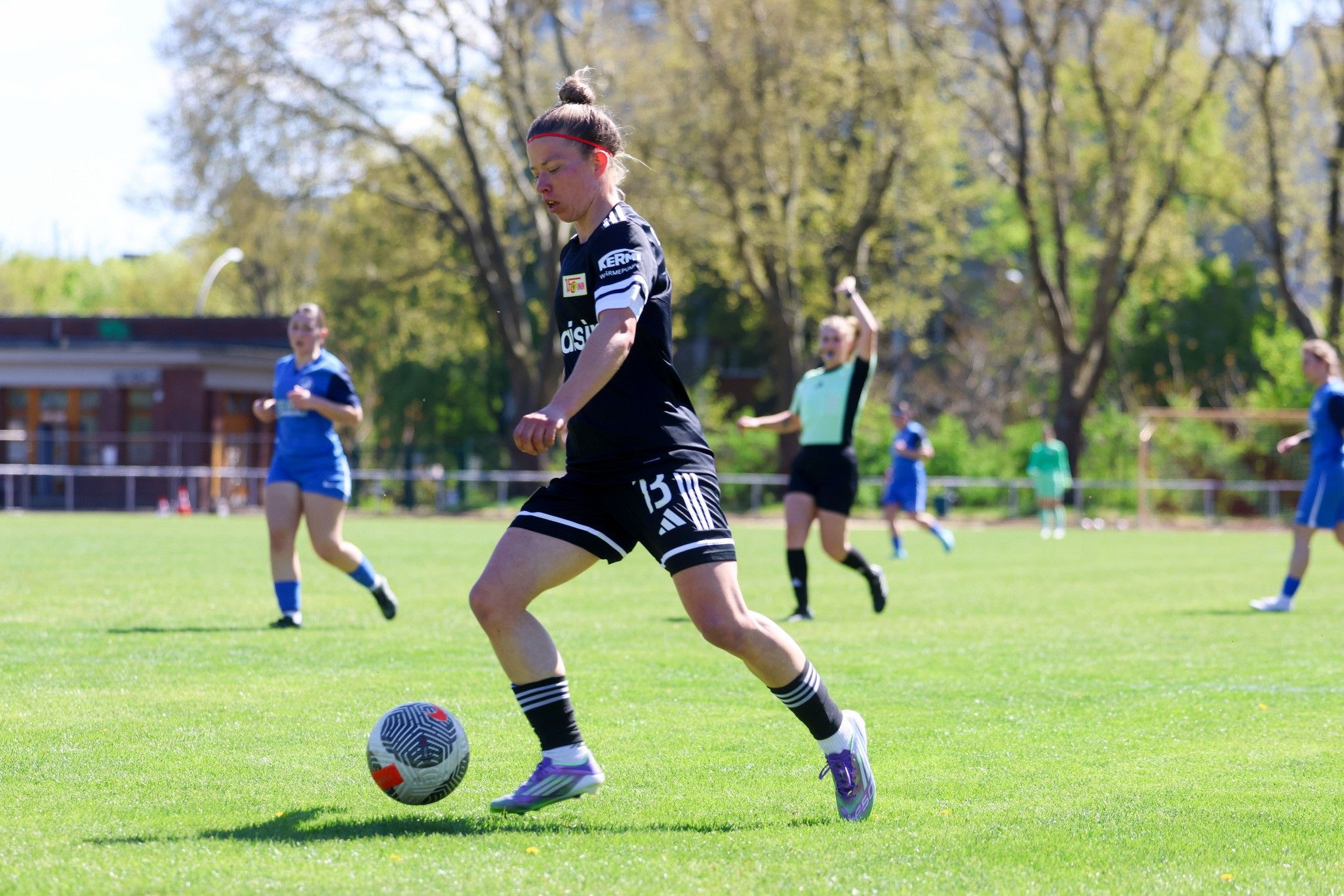 Eine Fußballspielerin in schwarz läuft mit dem Ball auf einem grünen Rasenplatz, während andere Spielerinnen im Hintergrund aktiv sind.