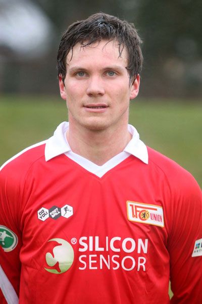 Ein junger Mann trägt ein rotes Fußballtrikot mit dem Logo eines Vereins und Sponsorenaufdrucken.