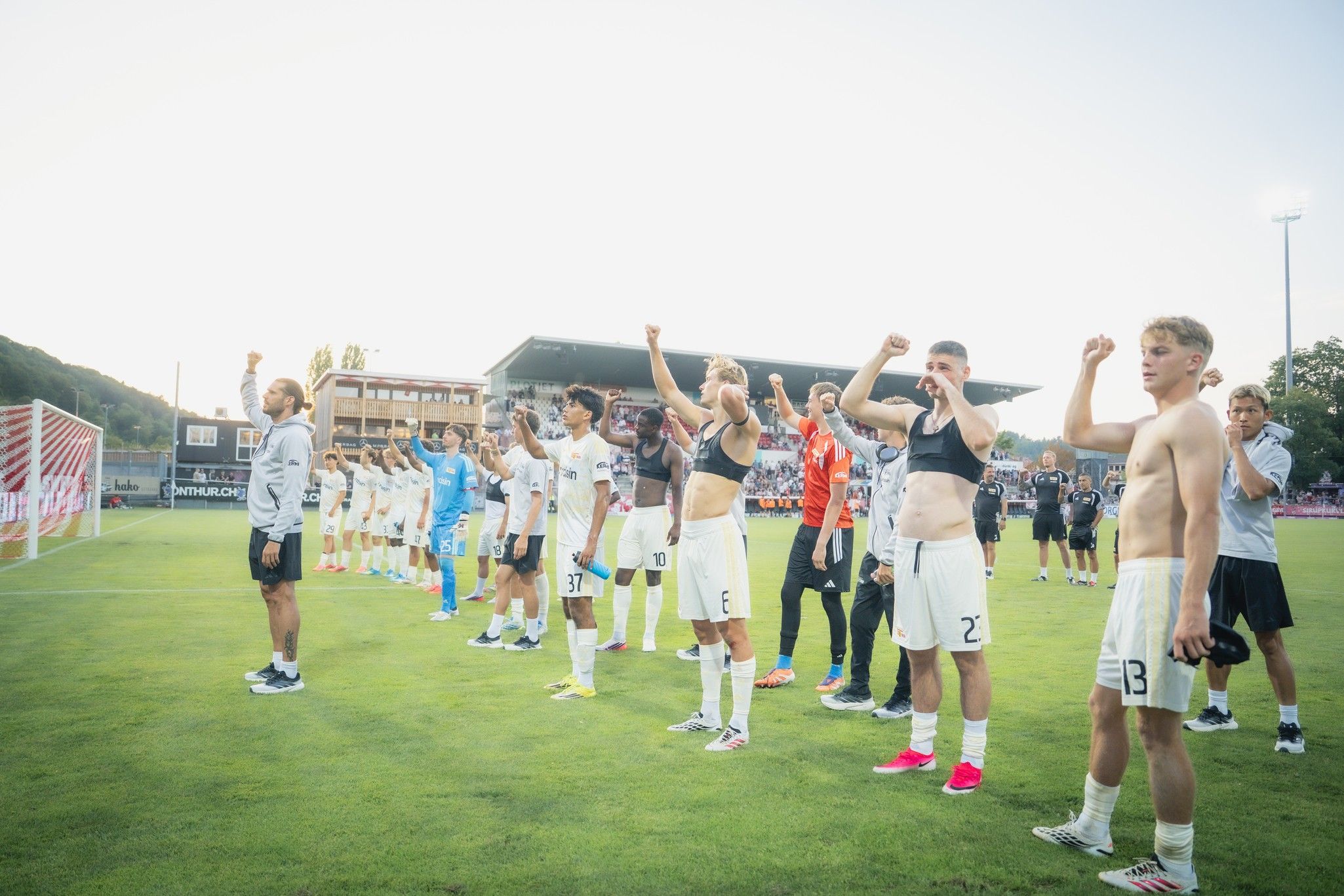 Fußballmannschaft steht auf dem Spielfeld, hebt die Fäuste und feiert, einige Spieler tragen Trikots, andere sind ohne Oberbekleidung.
