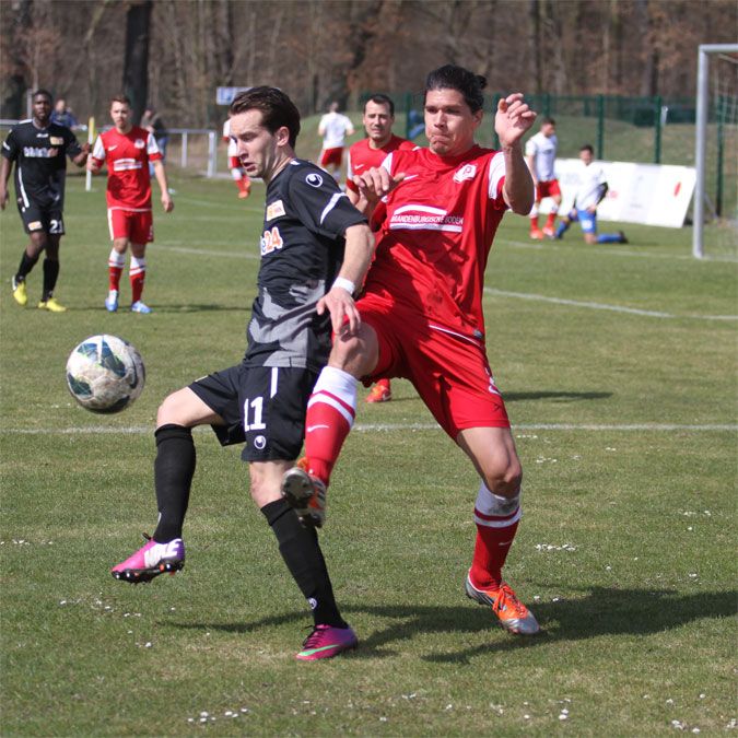 Zwei Fußballspieler in Aktion während eines Spiels, einer im schwarzen Trikot, der andere im roten Trikot, beide im Zweikampf um den Ball.