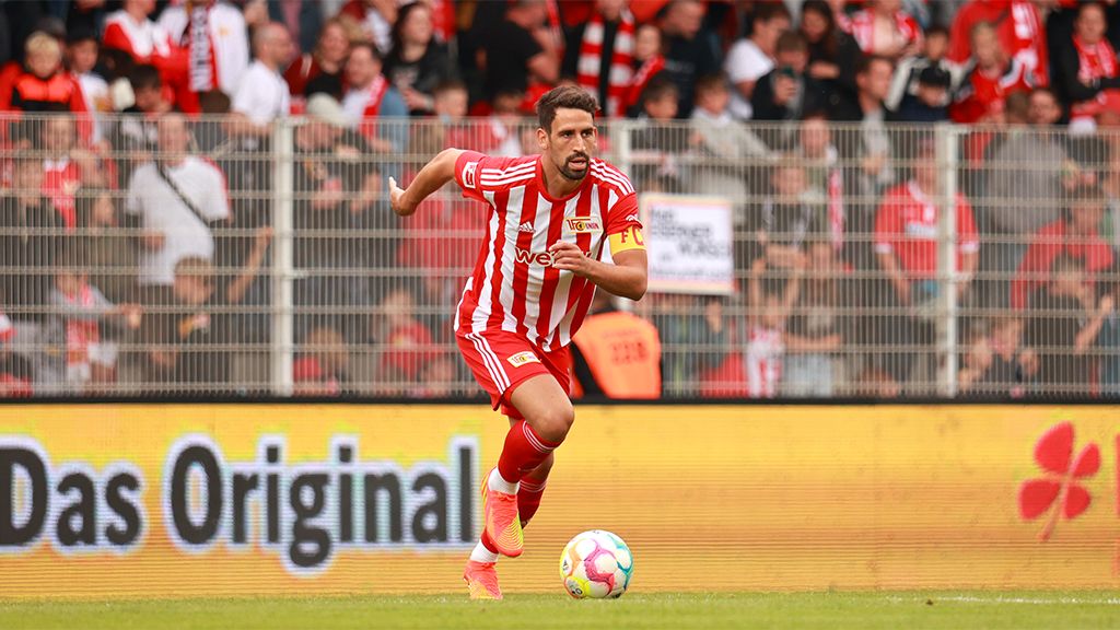 Ein Fußballspieler in rot-weißem Trikot sprintet über das Spielfeld, umgeben von jubelnden Fans im Hintergrund.