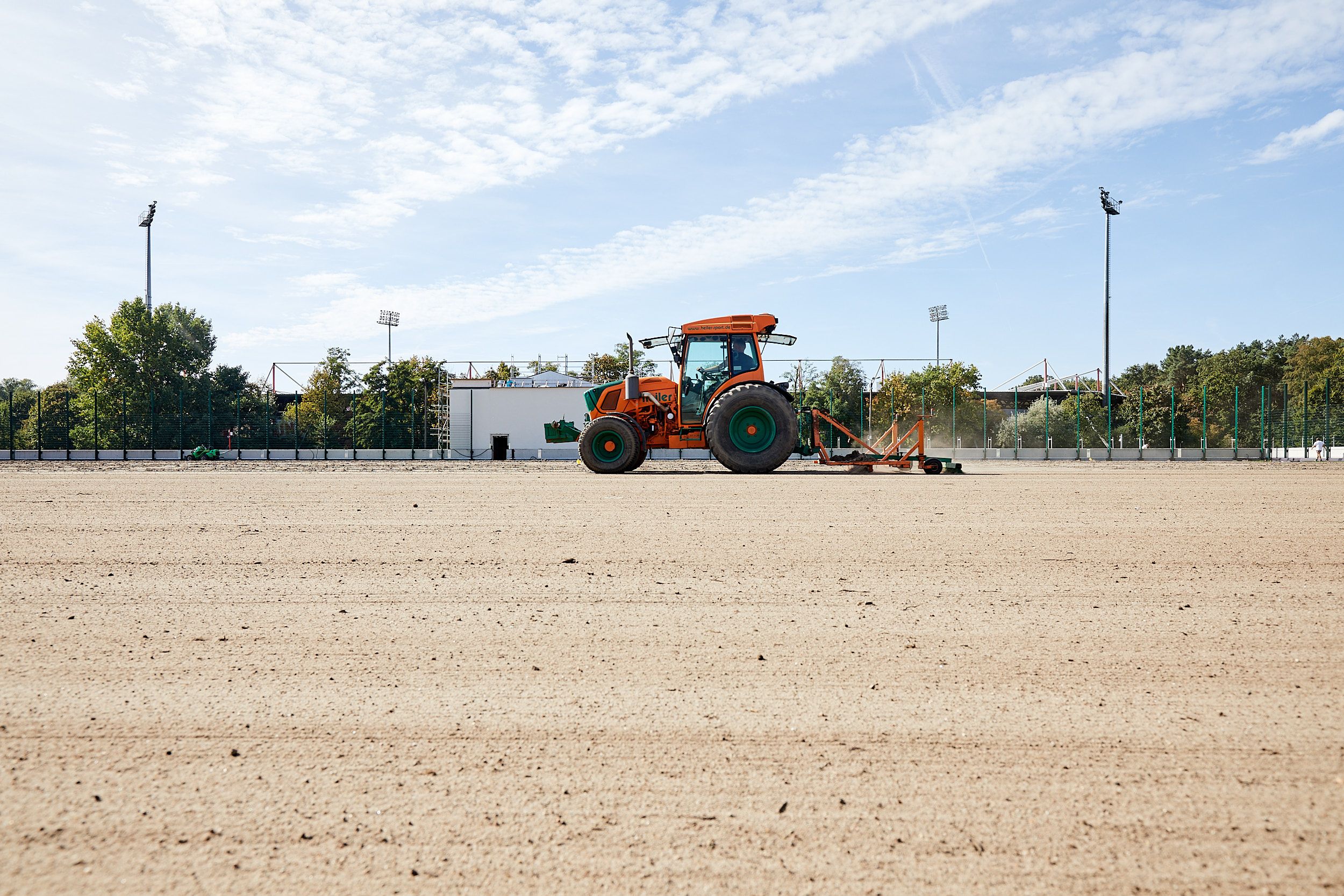 Ein orangefarbener Traktor bearbeitet einen trockenen, leeren Sportplatz im Freien bei sonnigem Wetter.