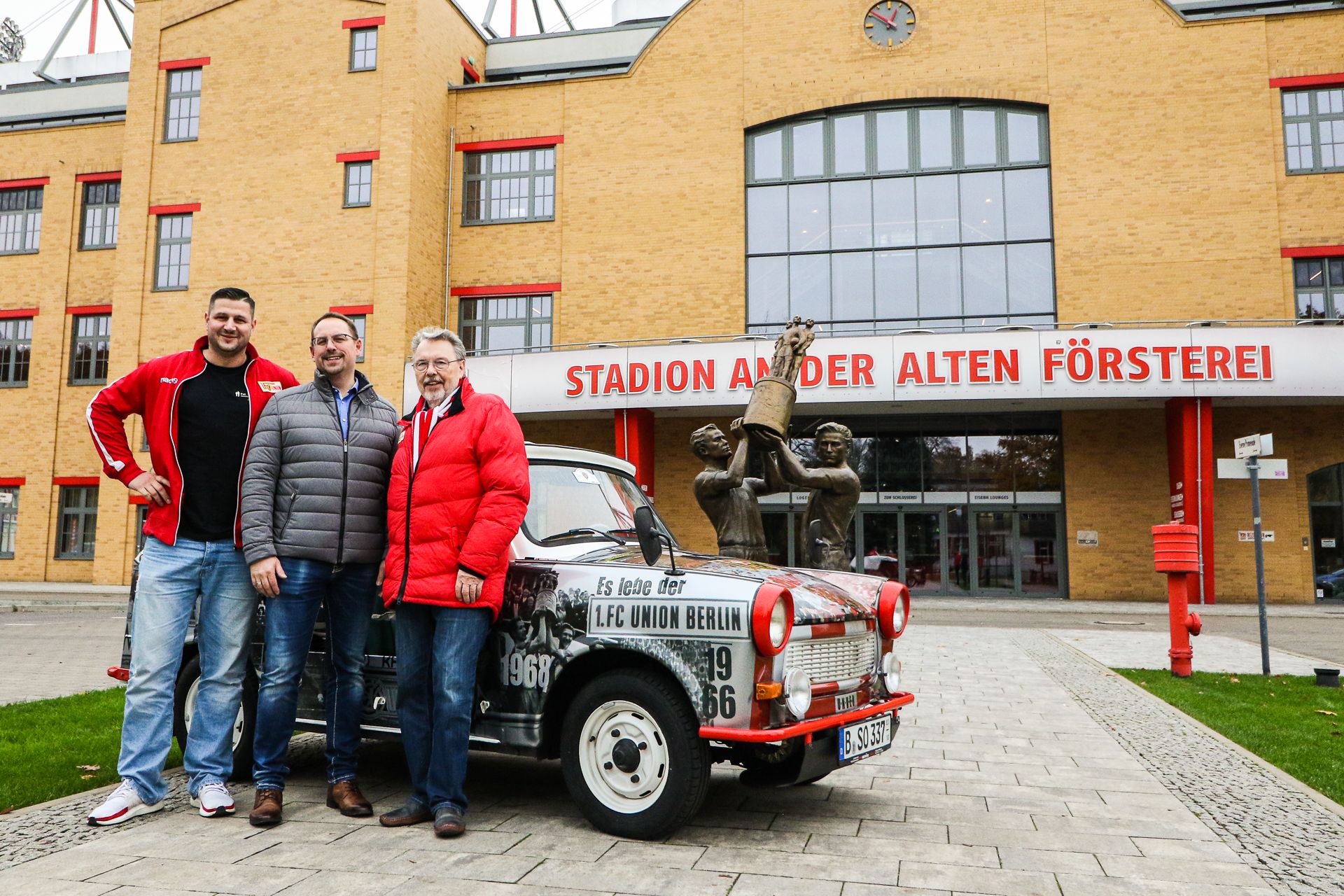 Drei Personen stehen vor dem Stadion An der Alten Försterei, neben einem typisch ostdeutschen Trabi-Auto.