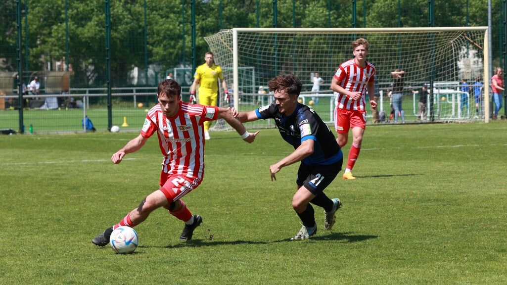 Zwei Fußballspieler im Wettkampf um den Ball auf einem grünen Spielfeld, im Hintergrund Zuschauer und weitere Spieler.