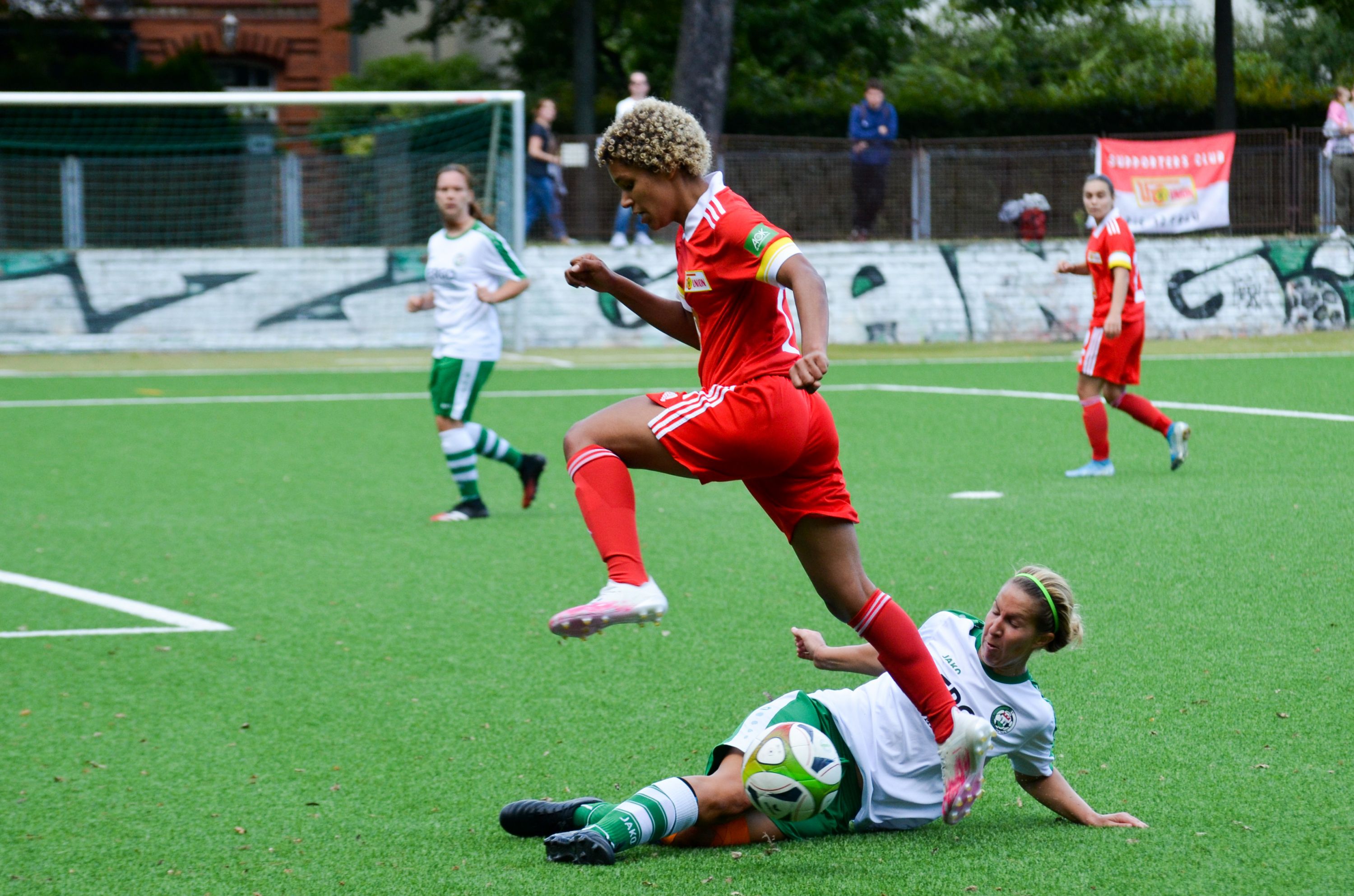 Eine Spielerin in rotem Fußballtrikot springt über eine liegengebliebene Spielerin in grünem Trikot auf einem Kunstrasenfeld.