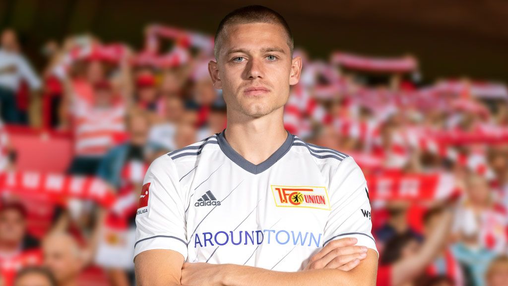 Ein Fußballspieler in einem weißen Trikot mit Vereinslogo steht vor einem Publikumsbereich mit Fans, die Schals halten.