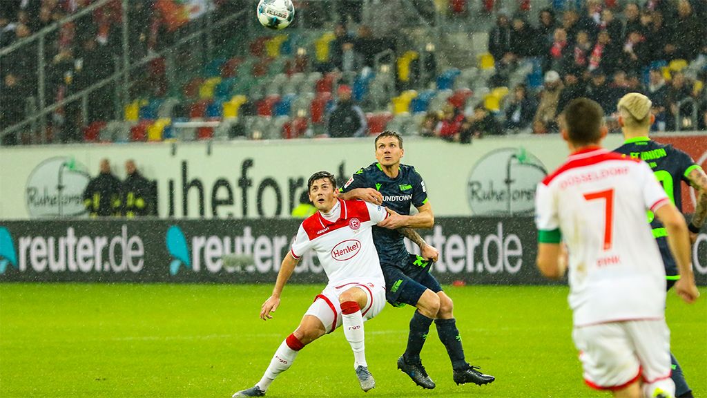 Ein Fußballspieler in rot-weißer Uniform hebt den Kopf, während ein Gegner in grüner Uniform den Ball nach oben spielt. Es regnet.