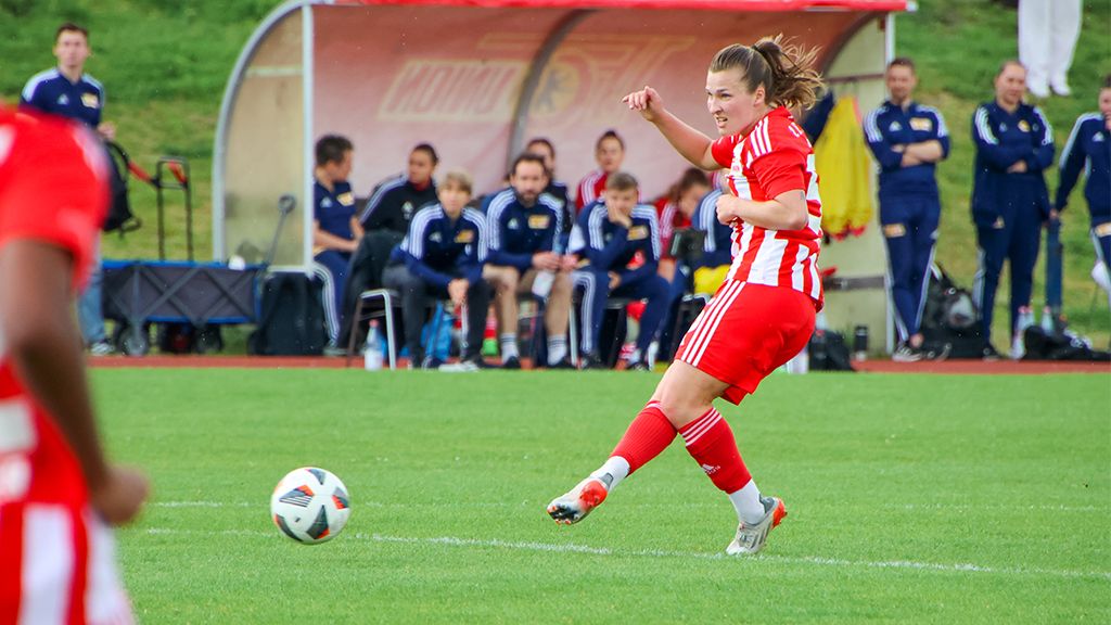 A female soccer player in a red jersey kicks the ball on a green field. Spectators sit in the background.