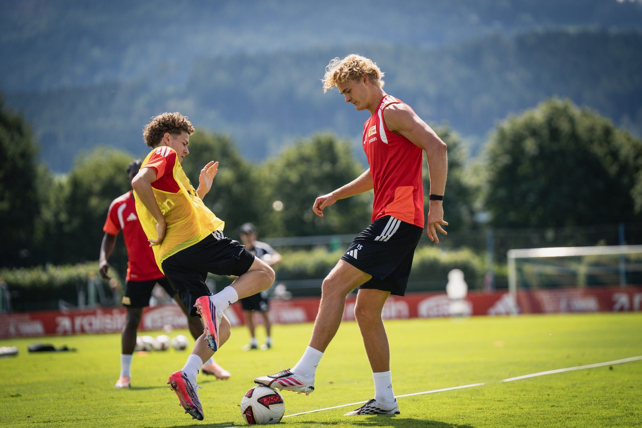 Zwei junge Fußballspieler trainieren auf einem grünen Platz, während andere Spieler im Hintergrund üben.
