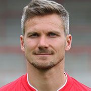 Porträt eines lächelnden Mannes mit grauen Haaren, in einem roten Trikot, vor unscharfem Hintergrund.