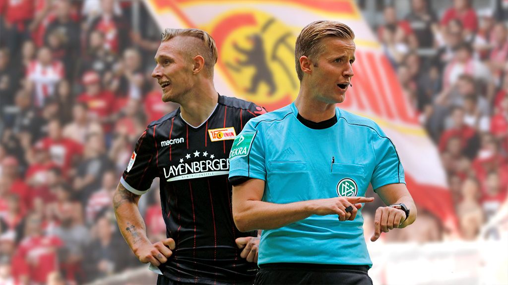 A football player in a black jersey stands next to a referee in a blue uniform, with a fan section in the background.
