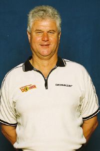 Ein lächelnder Mann mit grauen Haaren trägt ein weißes Poloshirt und steht vor einem blauen Hintergrund.