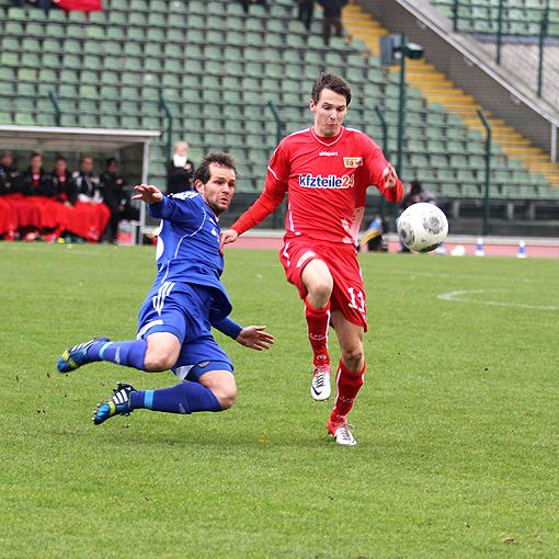 Zwei Fußballspieler kämpfen um den Ball; einer liegt am Boden, während der andere im roten Trikot den Ball tritt.