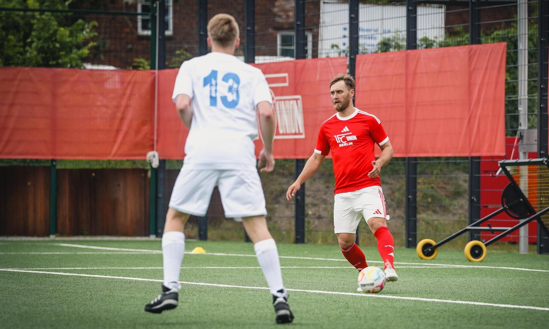 Ein Fußballspieler in rotem Trikot dribbelt den Ball auf einem Kunstrasenplatz, während ein Gegner in weißem Trikot ihn ansieht.