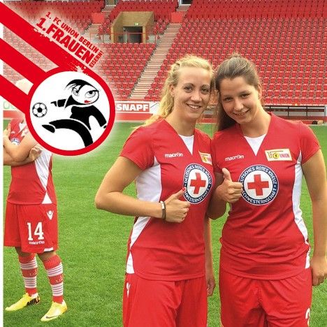 Zwei Fußballerinnen in roten Trikots geben Daumen hoch, im Hintergrund ein weiterer Spieler und ein Fußballstadion.