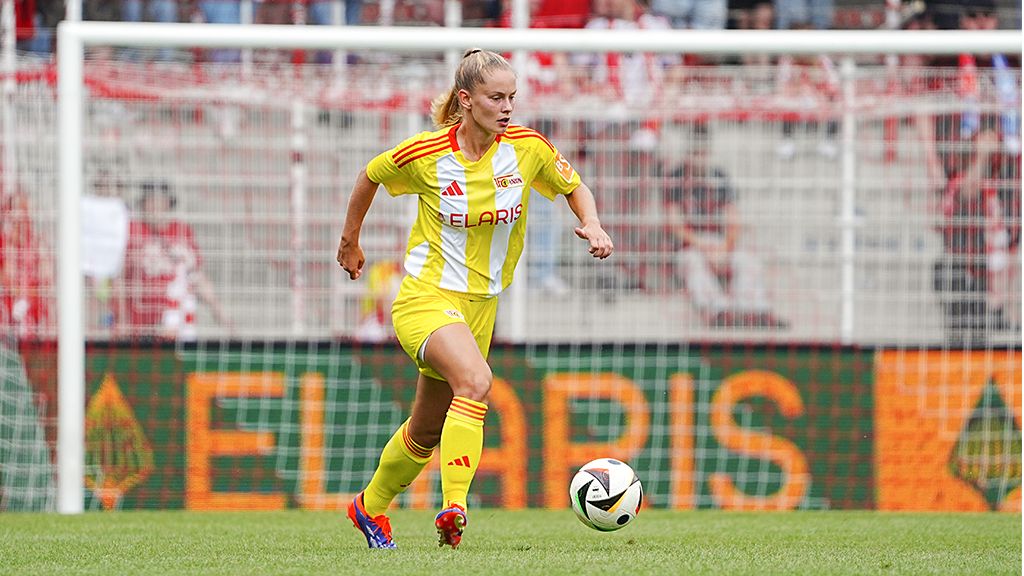 Eine Fußballspielerin in einem gelben Trikot sprintet über das Spielfeld, mit einem Tor im Hintergrund.