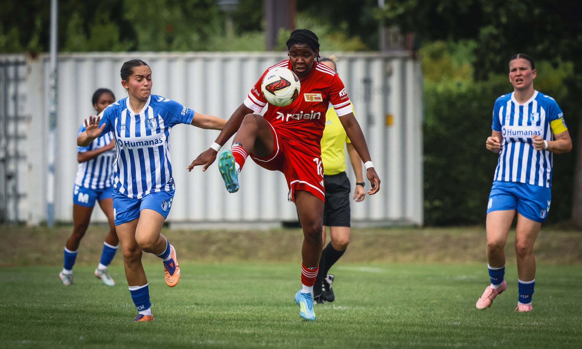 Eine Spielerin in rotem Trikot hebt den Fußball mit ihrem rechten Fuß an, während eine Gegenspielerin in blau-weißem Trikot folgt.