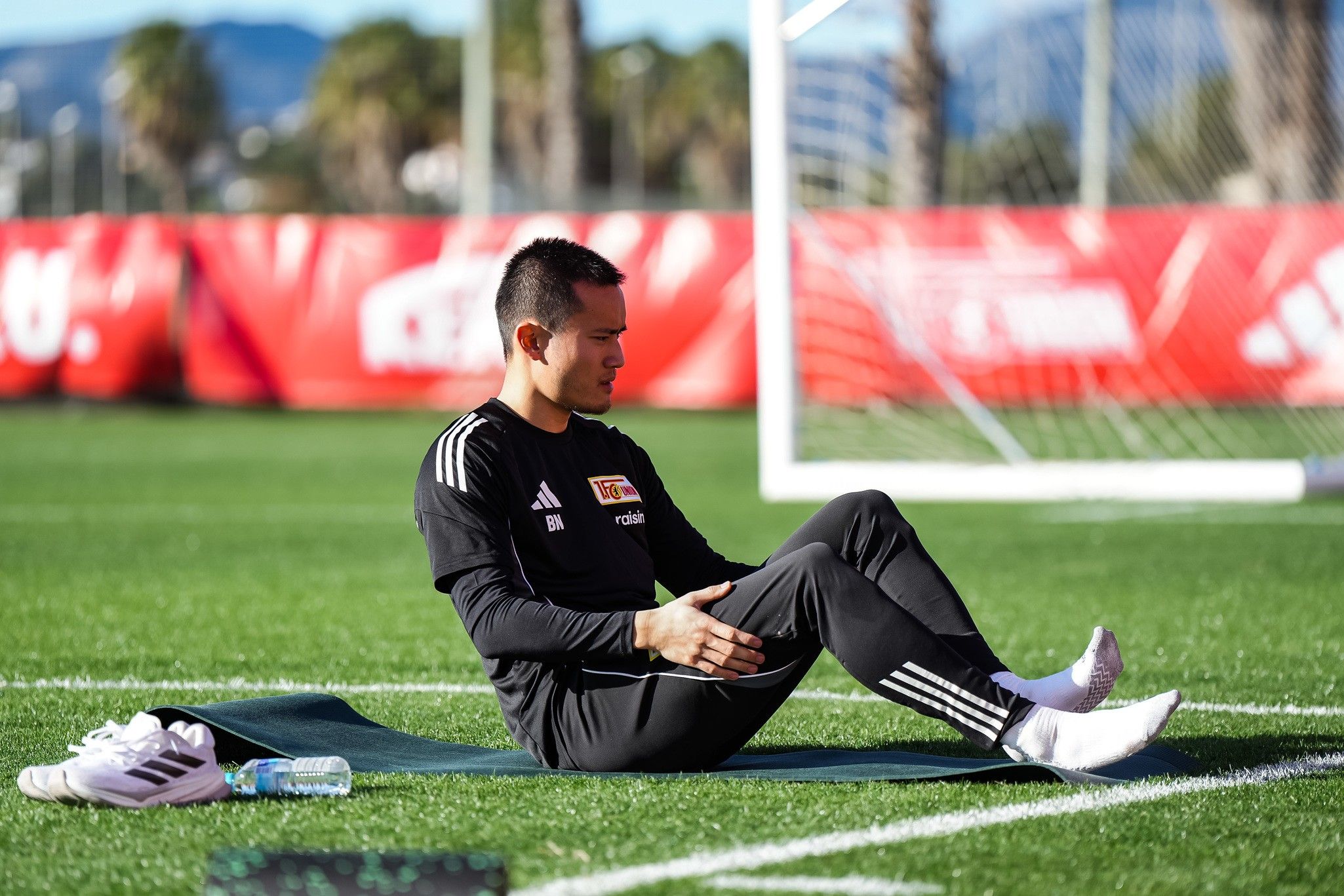 Ein Sportler sitzt auf einer Matte auf einem Trainingsfeld und führt eine Übung durch, im Hintergrund ein Fußballtor und Palmen.