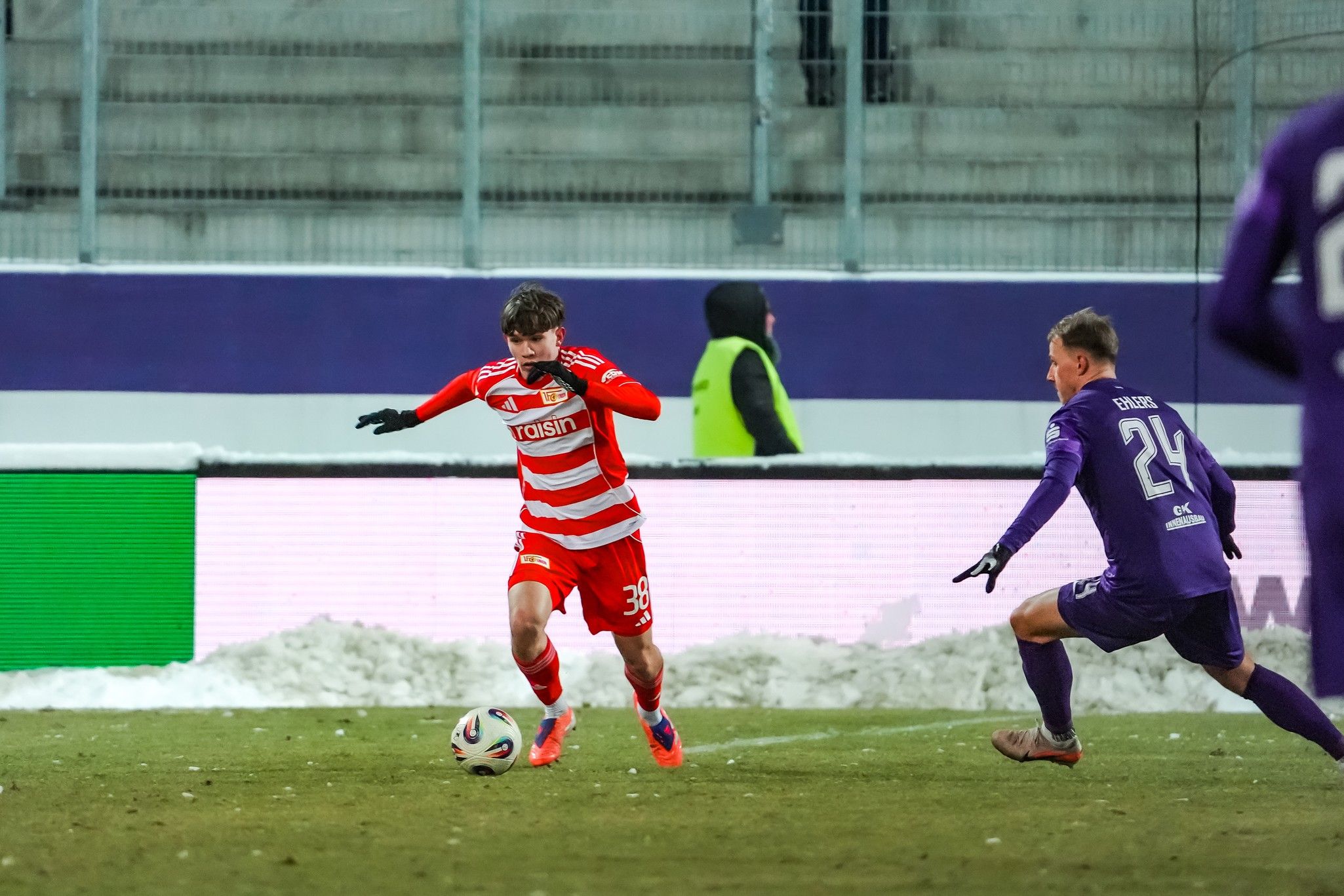 Spieler in rotem Trikot dribbelt auf dem Rasen, während ein Gegner in lila hinterherläuft. Schnee ist am Rand des Spielfelds sichtbar.