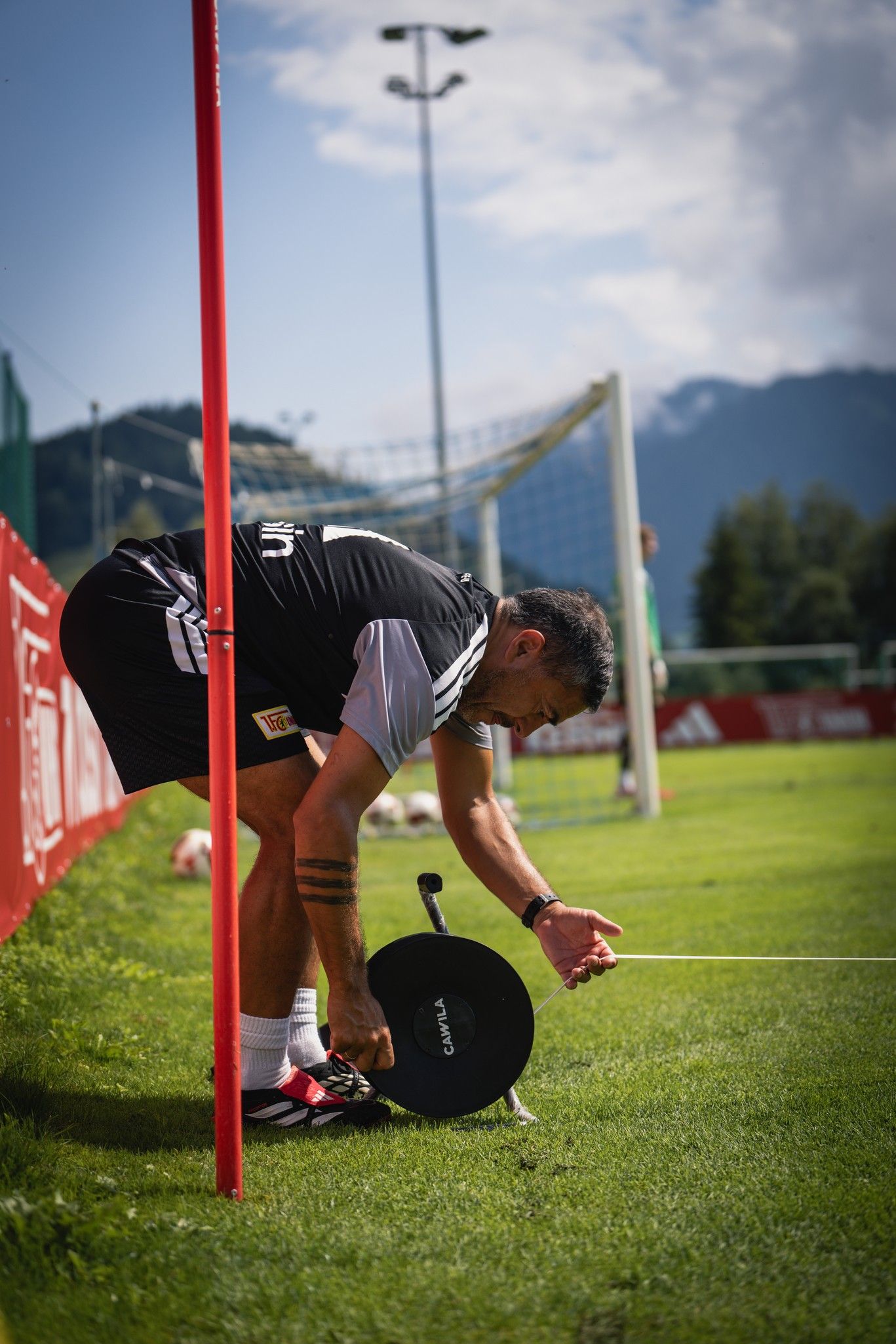 Ein Sportler justiert eine Trainingshilfe, während er sich an einem roten Torpfosten abstützt, im Hintergrund ein Fußballfeld.