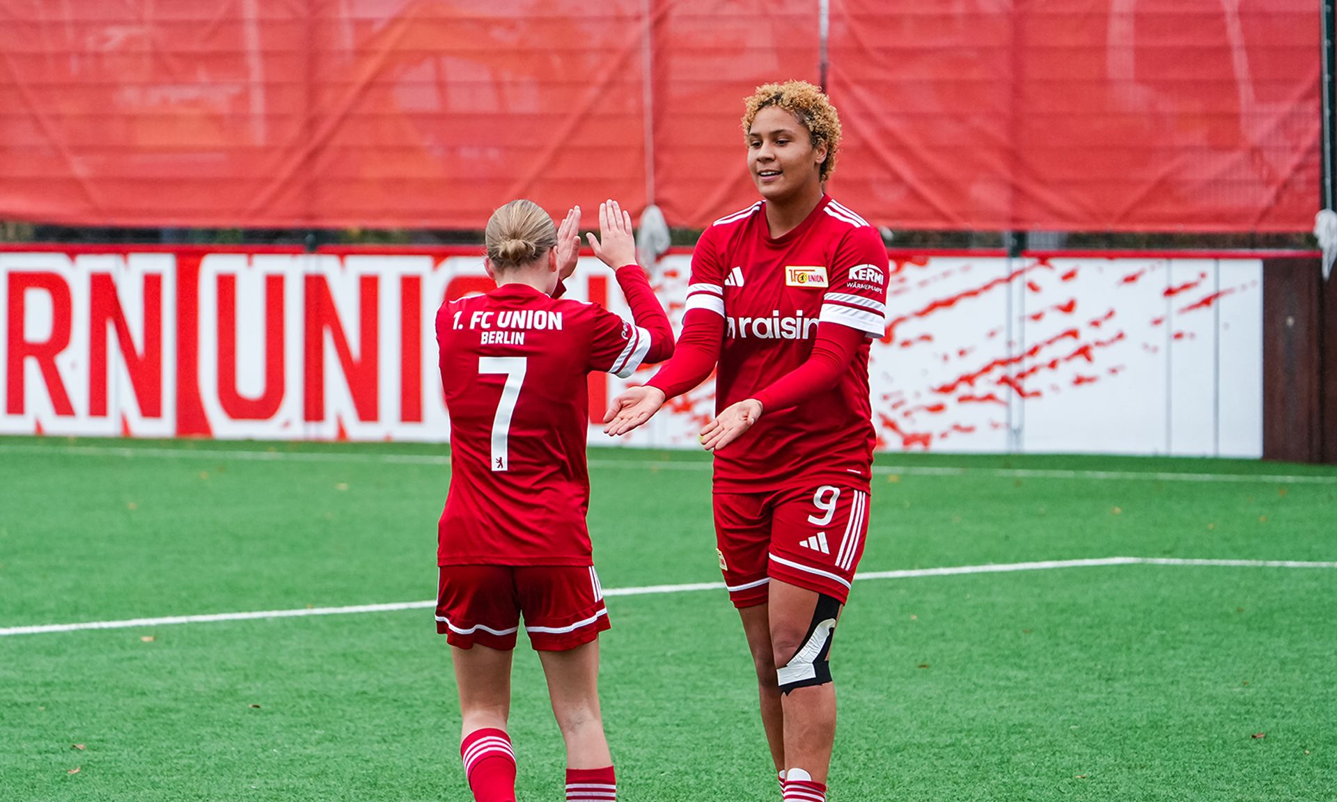 Zwei Frauen im roten Fußballtrikot stehen auf einem grünen Platz, lächeln und klatschen sich ab.