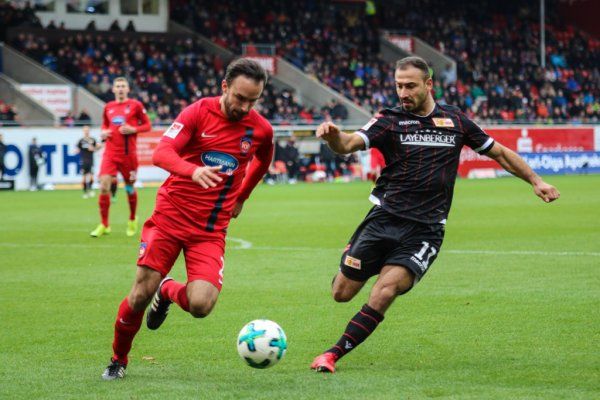 Two soccer players are battling for the ball during a game on a stadium field. Spectators can be seen in the background.