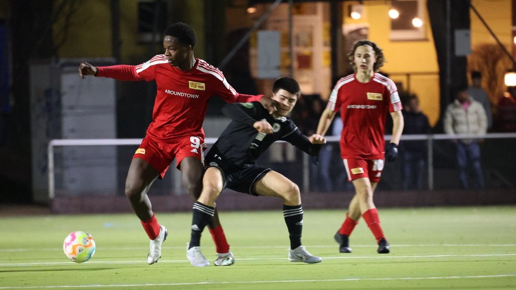 Ein Fußballspieler in rotem Trikot dribbelt, während ein anderer Spieler in schwarzem Trikot versucht, den Ball zu erobern.