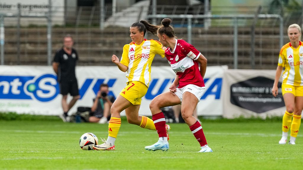 Two female soccer players in yellow and red jerseys are playing outside while a ball rolls between them.