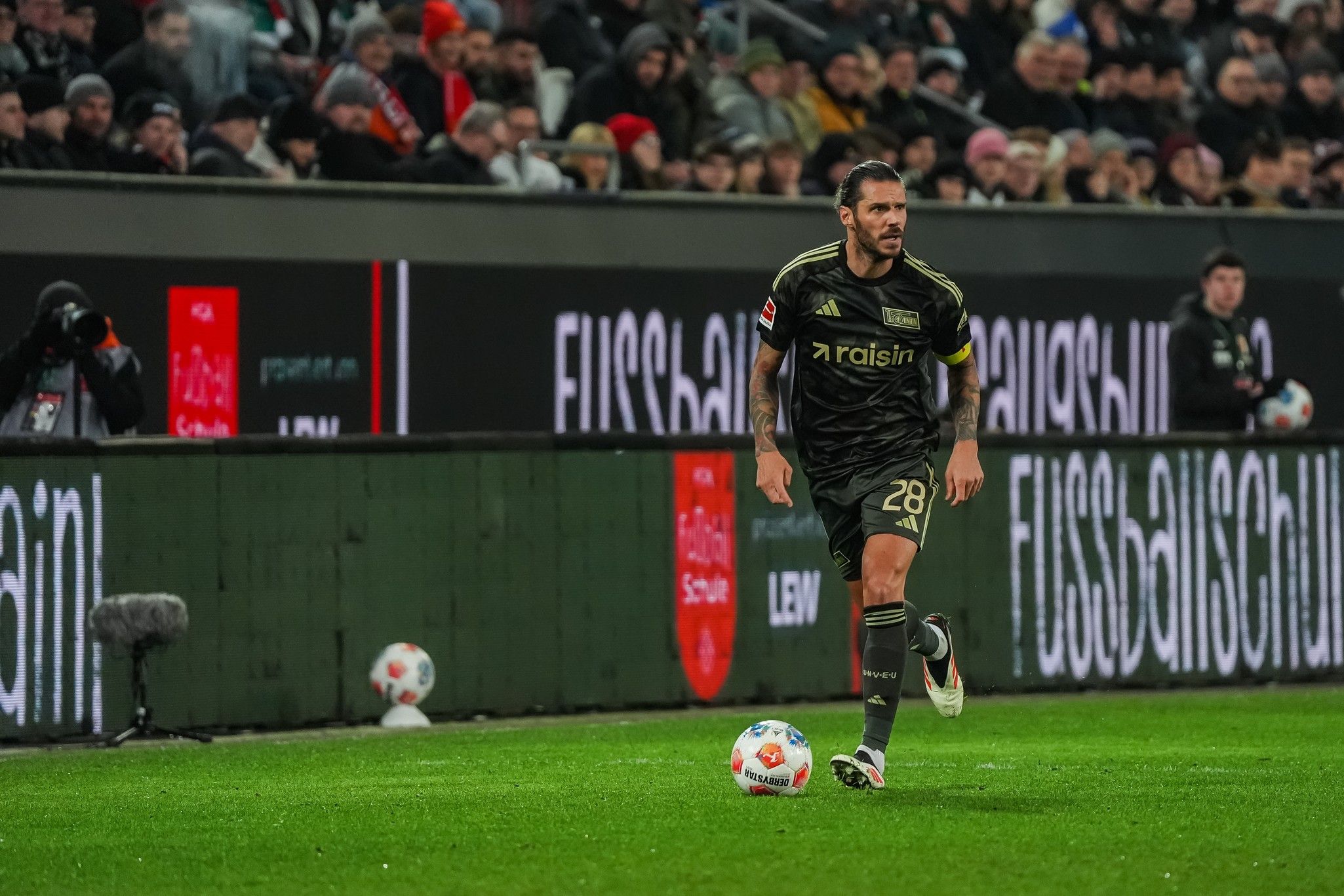 Fußballspieler in schwarzer Ausrüstung dribbelt den Ball auf einem Stadionfeld voller Zuschauer.