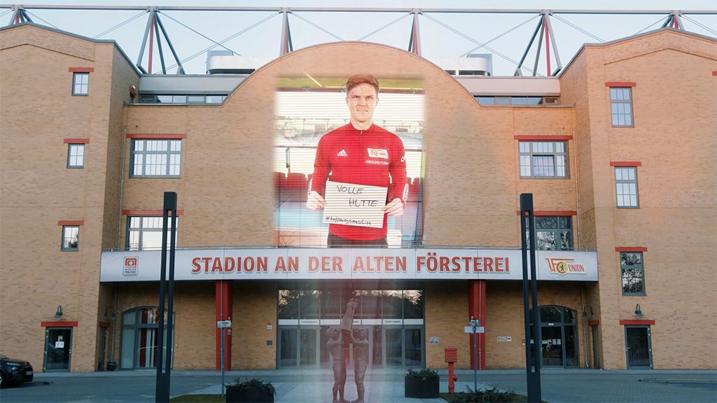 Ein Stadion mit roten und beigen Wänden, an dem ein Mann mit einem Schild steht. Über dem Eingang prangt der Schriftzug „STADION AN DER ALTEN FÖRSTEREI“.