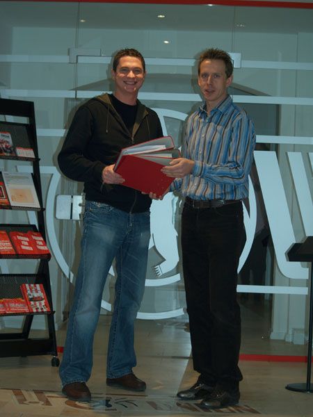 Zwei Männer stehen in einem modernen Büro, halten rote Ordner und lächeln sich an. Im Hintergrund sind Informationsmaterialien sichtbar.