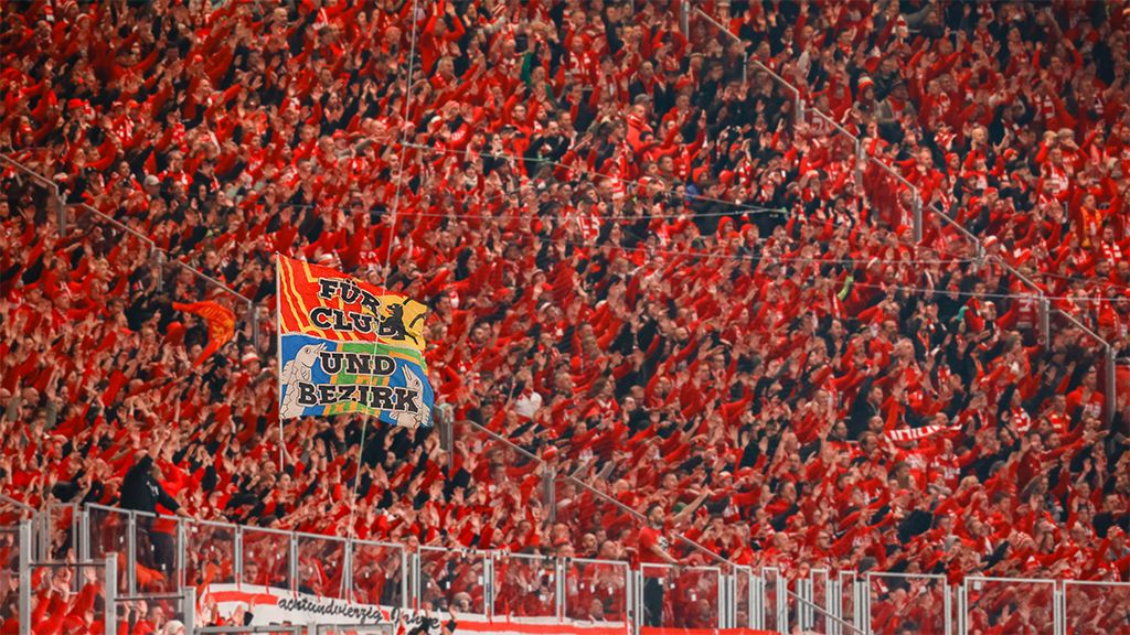 Ein begeistertes Publikum in roten Kleidern jubelt, während ein Banner mit der Aufschrift „FUSSBALL CLUB“ und „UND BEZIRK“ geschwenkt wird.