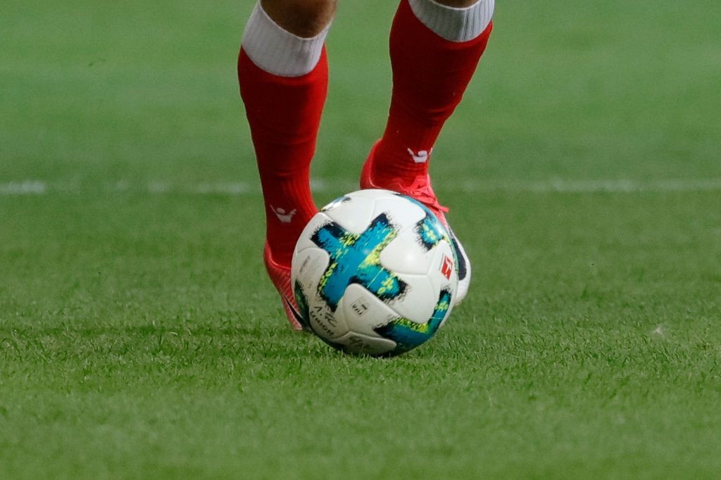 Spielerfüße in roten Socken berühren einen Fußball auf einem grünen Rasenplatz.