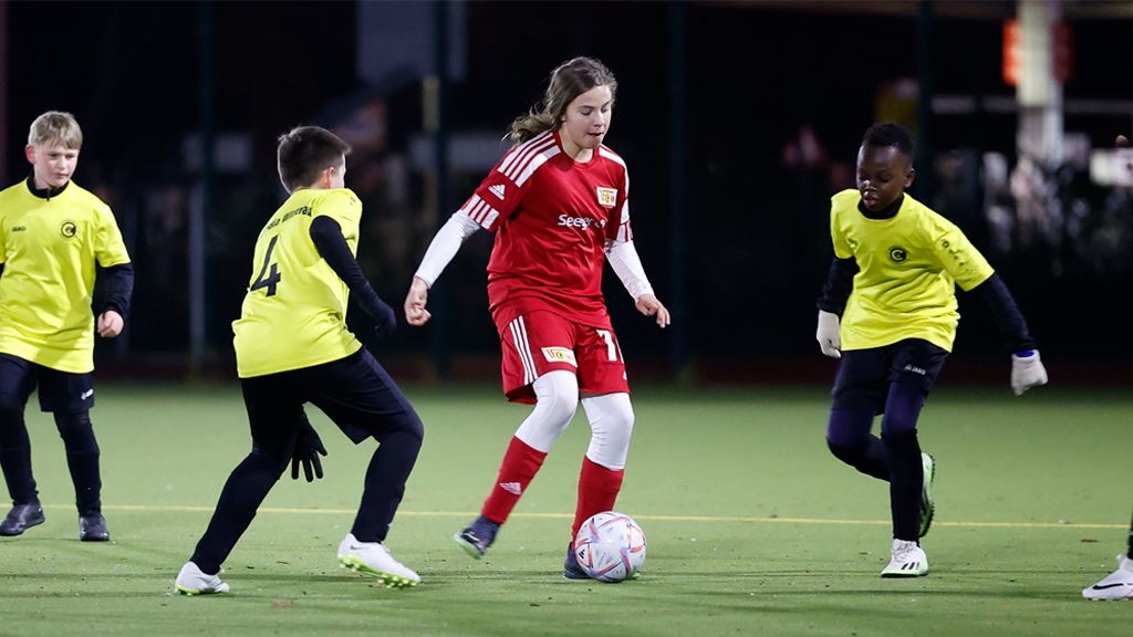 Mädchen in rotem Trikot dribbelt den Ball um zwei Spieler in gelben T-Shirts auf einem Fußballplatz bei Nacht.