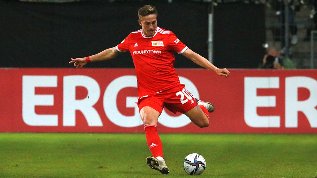 A player in a red jersey kicks the ball during a soccer game. The background shows advertising banners.