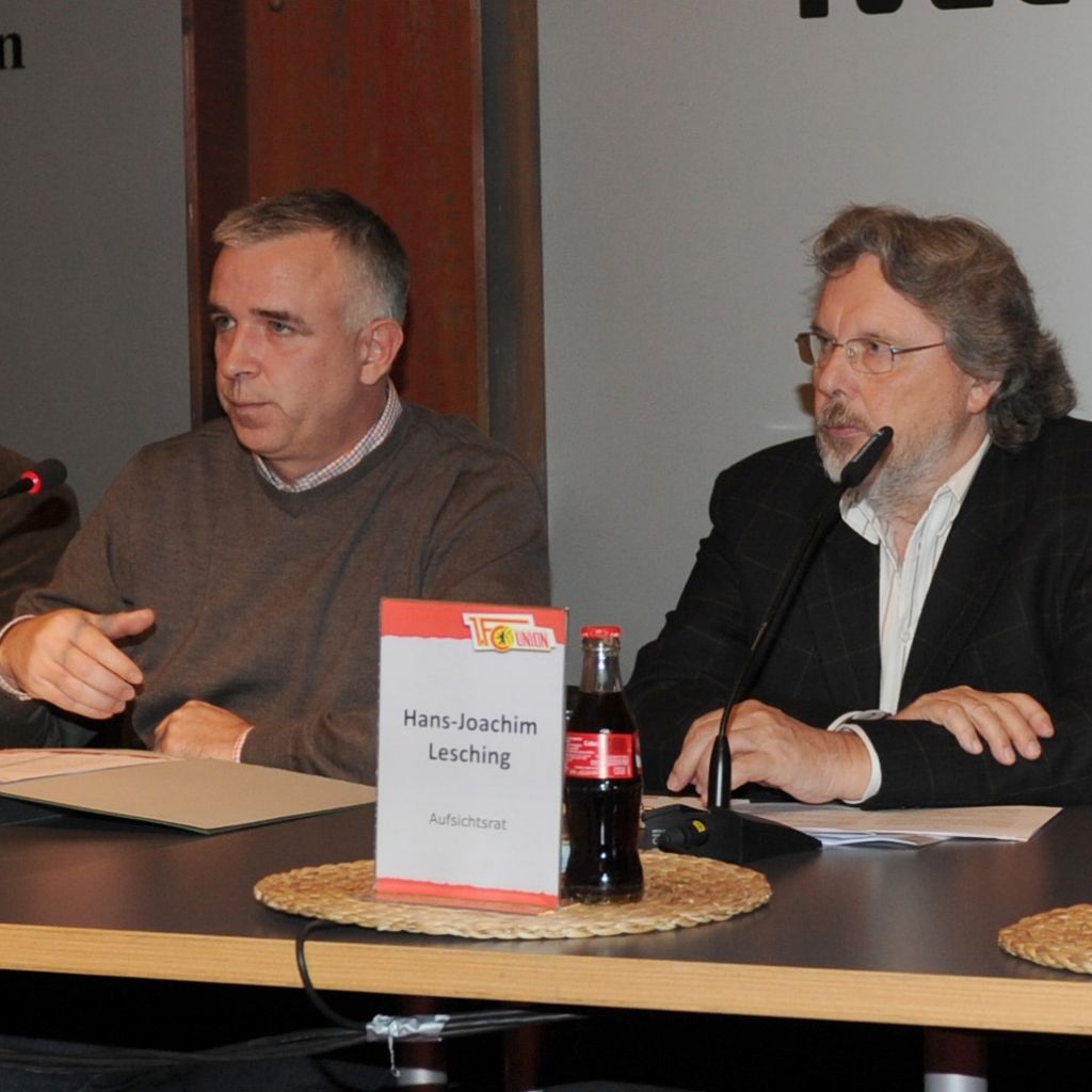 Zwei Männer sitzen an einem Tisch mit Mikrofonen, im Hintergrund ein Schild mit dem Namen Hans-Joachim Lesching.