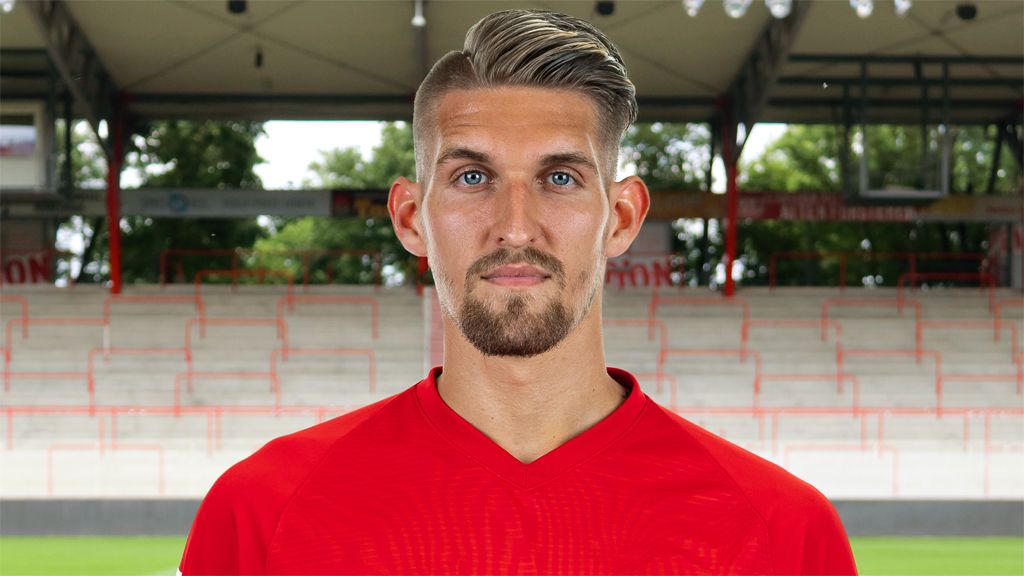 Ein junger Mann mit kurzen, blonden Haaren und Bart trägt ein rotes Trikot vor einem leeren Stadionhintergrund.